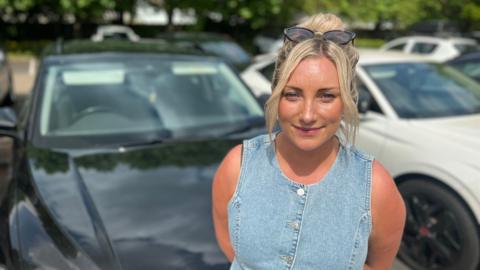 Sophie Rose, who has blonde hair tied up and sunglasses on her head, wearing a denim sleeveless top and standing in front of cars in a car park while looking into camera