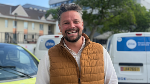 A man stands outdoors in front of parked electric vehicles marked with the EVIe logo, wearing a brown padded vest over a light‑coloured shirt. Residential buildings, trees, and a street are visible in the background.