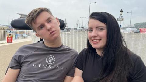 An image of Jack Dolan and mum Lisa Dolan in Dover, Kent. Lisa has long black hair and is wearing a black t-shirt. Jack, who is in a wheelchair, is wearing a grey 'Gymking' t-shirt. 