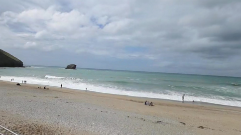The picture shows Portreath beach which has a mix of sand and pebbles. Several people are visible, some standing near the water, others on the sand. The ocean has gentle waves, and a large rock formation is visible in the water near the horizon. 