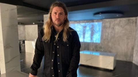 Long haired man Michael O'Hare wears a dark shirt and looks at the camera in a restaurant whose walls appear to be made of granite