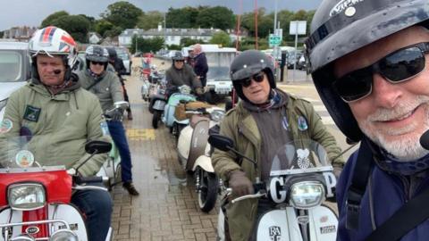 The image is a selfie taken by a man in a helmet, it shows him smiling at the camera and behind him are a group of men on scooters in two lines parked one behind the other on a road. 
