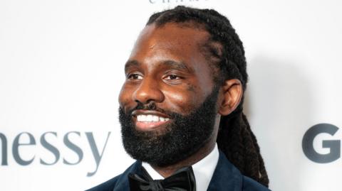 Wretch 32 at the GRM Gala. Wretch has long braided hair tied back and a short beard. He smiles, looking to the left of the camera, while wearing a black suit with velvet bow tie