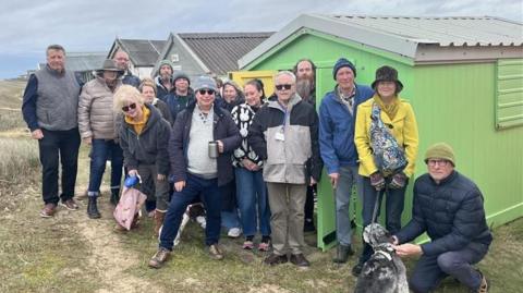 A group of beach hut owners standing outside a row of brightly coloured wooden properties.