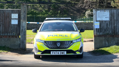 A marked fluorescent yellow and blue police car is parked outside of one of the entrances to Sparrow's Nest Gardens. Behind the car, which is parked, there is a blue and white police tape.