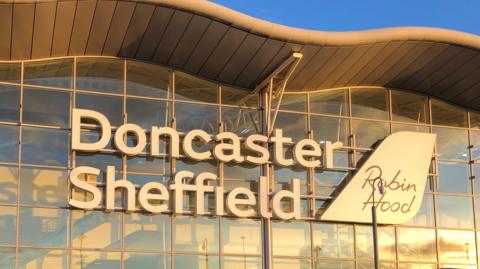 The outside of the former Doncaster Sheffield Airport building is covered in windows, it has its name in large white letters and the words Robin Hood on a sign shaped like the tail of a plane