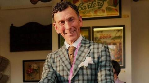 A photo of Matthew Upham looking at the camera. He is wearing a grey checked jacket, a light brown tie and a pink lanyard