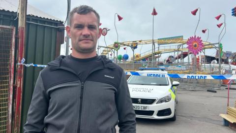 John, who has short brown hair and stubble, wearing a black and grey hoodie. There is a police car behind him in front of a police cordon. In the background the amusement park can be seen with a rollercoaster track fenced off by metal gates.