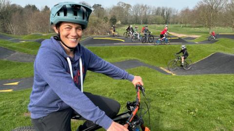 A woman wearing a mid-blue hoodie and a turquoise cycle helmet sits on a bike and smiles at the camera. In the background a number of children are riding bikes on a pump track, a bike track with lots of hills of various sizes.