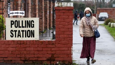 Woman wearing a coat with her hood up walks on a pavement towards the camera, past a sign that reads: "Polling Station"