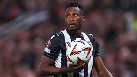 Baba Rahman, wearing a black and white striped PAOK shirt, holds a football in the palm of his right hand as he turns his head to his right over