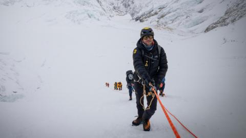 Tim Howell is climbing up a mountain holding on to rope. He is wearing weather protective gear. There is a team of people behind him.