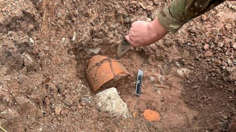 Somebody digging up artefacts. A terracotta pot can be seen in the ground next to a measuring ruler. 