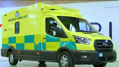 The photo shows an ambulance which is parked in a garage. It's shell has been painted with the distinctive yellow and green painting. It reads 'NHS Emergency Services' on the side. 