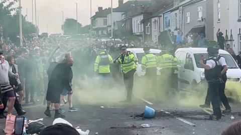 A street is filled with people and various police officers are stood around holding batons. Smashed items can been on the tarmac of the road and there is some smoke circling. 