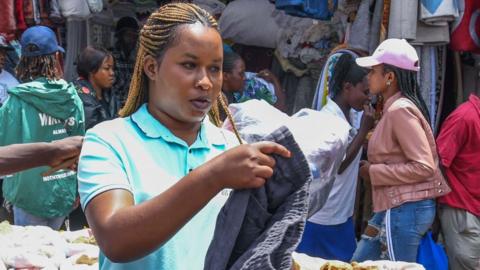 A clothes vendor with long braids and in a blue T-shirt (L) holds up black trousers at Gikomba market in Nairobi, Kenya. Behind her can be seen people walking past other stalls -September 2023.