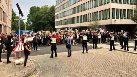 A line of police officers with protesters facing them some holding placards