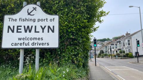 A picture of Newlyn. The sign on the left reads "The fishing port Newlyn welcomes careful drivers". On the right is the road and traffic lights. 