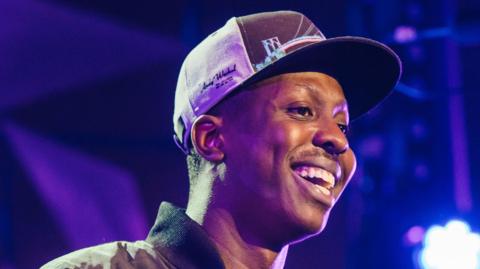 A photo of Jamal Edwards wearing a cap and smiling. It is against a backdrop that looks like a stage. 