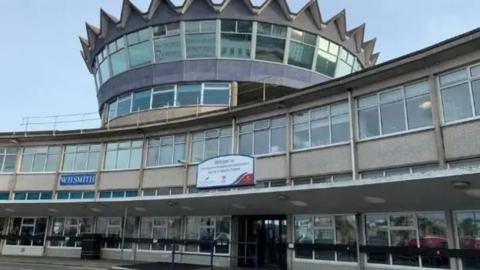 The exterior of the sea terminal building a two storrey brown building with a round turret shaped third storrey in the middle with a spikey roof, there are large windows along the structure.