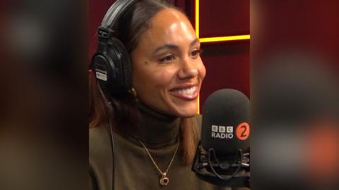 Alex Scott has long brown hair and wears a brown turtle neck. She has headphones on and is in a radio studio