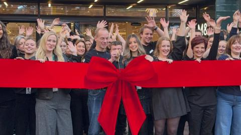 A crowd of shoppers all waving with their hands in the air and smiling at the camera. They are standing behind a giant red ribbon with a big red bow which is about to be cut. The doors to a Marks and Spencer store is just visible behind them.