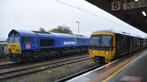 Trains on the tracks at a rail station on what appears to be early morning. The signing on one of the trains reads Maritime. The ground is wet.