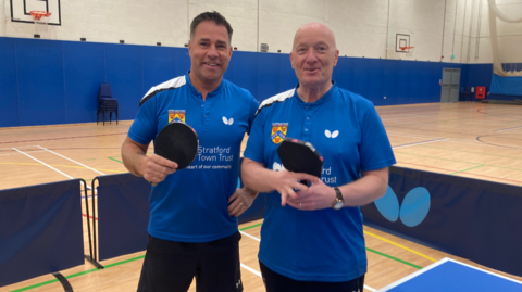 Two men, each in blue tops and holding table tennis bats, stand in a sports hall smiling at the camera