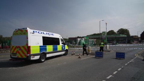 Police officers standing guard next to police tape, with a petrol garage in the background. A police van is also parked on the road.