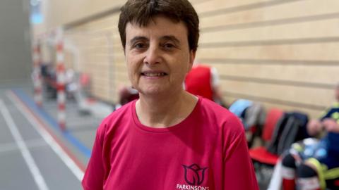 Julie Norris is a woman in her sixties wearing a pink top. She has dark hair and is smiling 