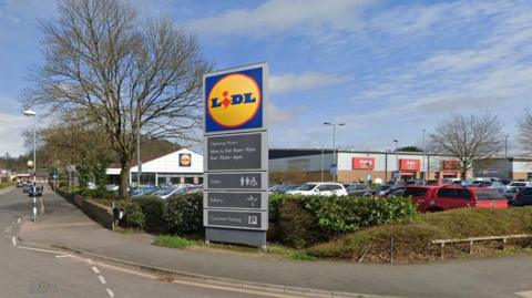 A junction of a road with a lidl sign face on which is in front of a Lidl car park.