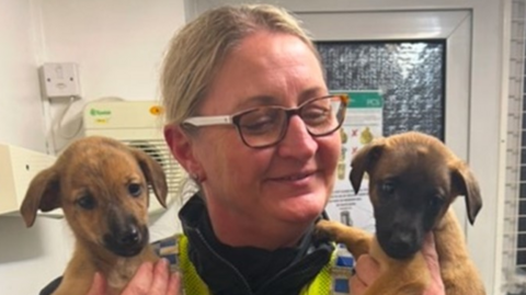 PC Kate Atkinson, who is wearing a police uniform and has blonde hair and tortoise shell glasses, holding up two of the lurchers that have been found.