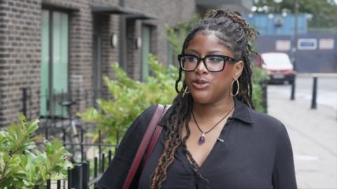Woman with dreadlocks, black shirt, and purple necklace standing on a sidewalk near a brick building and greenery.