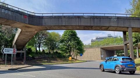 A blue car travels around the Five Bridges roundabout. The roundabout's concrete bridges stand above it, and splits off into multiple directions. The Gateshead flyover can be seen in the background.