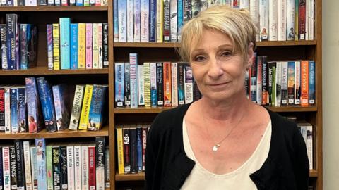 Jennie Storey wearing a white dress, black cardigan and appearing with dyed blonde hair, stands in front of shelves full of books. She is looking at the camera