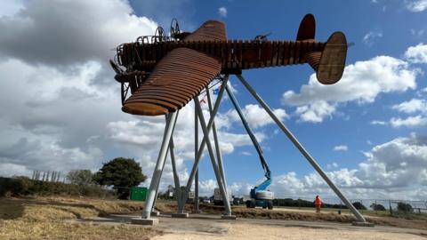 A life-size steel brown sculpture of a Lancaster bomber in a flying motion and tilted to the side. It is held up high by several posts and construction vehicles and a person in orange hi-vis can be seen next to it.