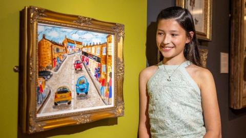 Sophia is standing beside her painting which is hung on a mustard yellow wall. She has long brown hair, is smiling, and is wearing a white dress. The painting is of cars driving up a road. Either side of the road are bright vibrant buildings.