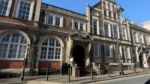 The exterior of the Douglas Council building, which is a large grand grey-brown building with large arched windows and steps up to a arched entrance.