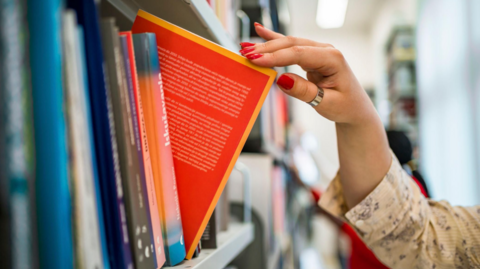A hand is holding a red and orange book, which is partially out of a shelf and at an angle to other books on the shelf.