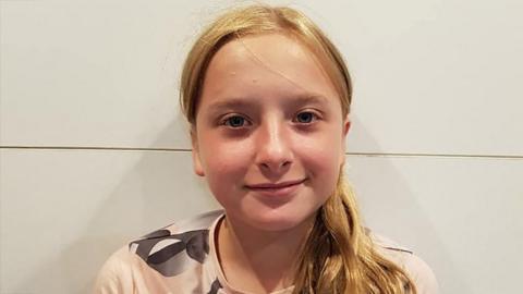 Lola Daviet is photographed standing against a white wall. She has blonde hair in a pony tail and is wearing a light pink t-shirt with grey and white detailing on the shoulder. 