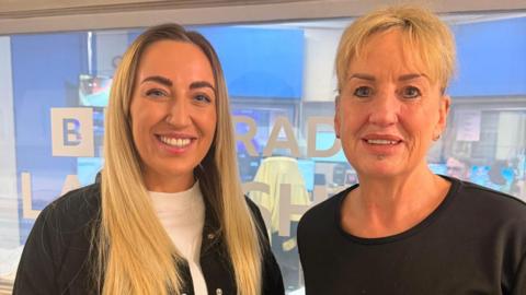Becky Hobkirk has long blonde hair a white sweater and black jacket. Mum Nicky has shorter blonde hair and a black top. Both are smiling in front of BBC Radio Lancashire glass door. 