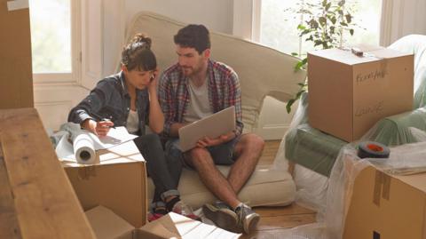 A man and a woman sit on a cushion looking at a laptop computer surrounded by unpacked boxes.