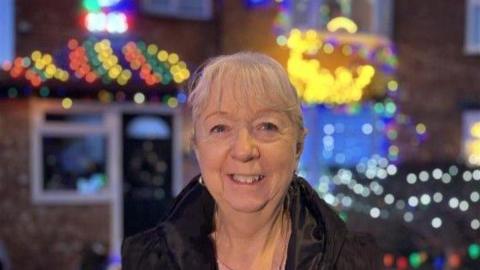 Irene Philpot wears a dark jacket and smiles into the camera in front of her home, which is decorated for Christmas.