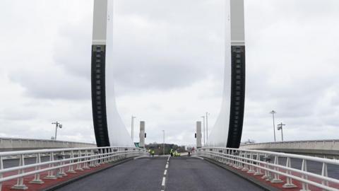 A view of the single-carriageway road that carries traffic in both directions over the Gull Wing bridge. There are barriers either side of the road and a pavement beyond them. Two large white and black "wings" point upwards from the middle of the bridge.