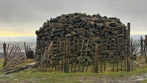 Large hut-size cairn made of large stones and surrounded by a circle of wooden fencing, which has partially fallen. It stands at the top of a hill, with flatter land spread out below.
