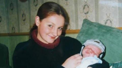 Picture of Paula Barrett with dark hair and wearing a blue sweater holding a baby dressed in light blue