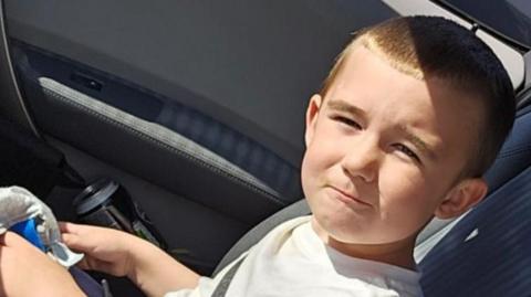 Lincoln Button is sitting in a car. He is squinting into the sun as he poses for the camera. He has cropped brown hair and is wearing a white T-shirt.