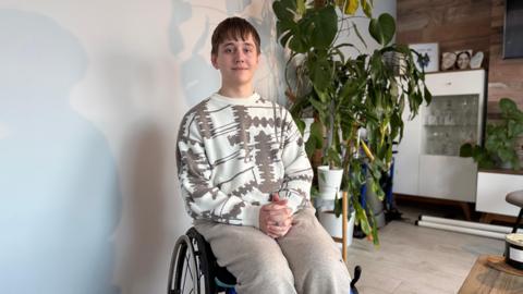 A teenage boy is in a wheelchair in a house. He is wearing a cream coloured jumper and grey trousers and is smiling. There is a large houseplant behind him.