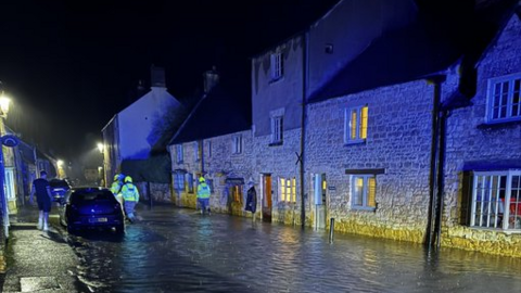 Flooding in Chipping Campden. The picture shows a residential street. There are crews in high vis jackets wading through the floodwater. Cars can be seen parked on the side of the road. 