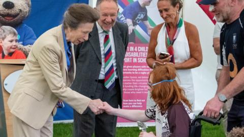 Princess Anne, the Princess Royal makes a presentation to a player in a wheelchair at Tag Fest 26. The Princess is wearing as a buff-coloured trouser suit over a blue blouse. The player shaking hands with her is in a wheelchair. They have red hair and glasses that are held on with a plastic band around their head.